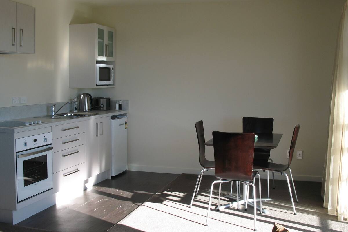 A modern kitchen with a built-in microwave, oven, and kettle, a dining area with a table and chairs, and a window with curtains.