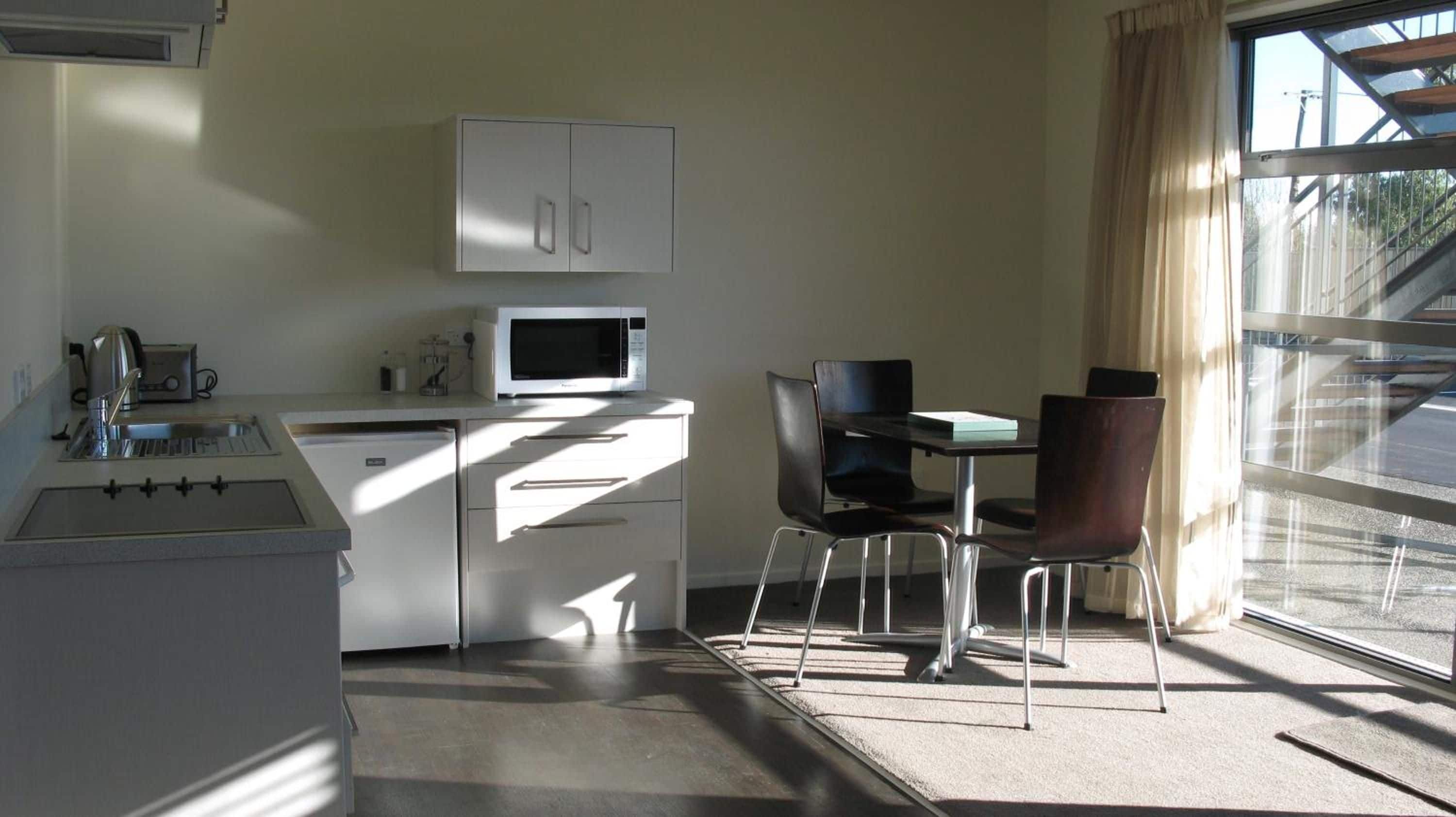 A compact kitchenette with a microwave, a sink, and a kettle, a small dining table with chairs, and a large window with a view of the outside.