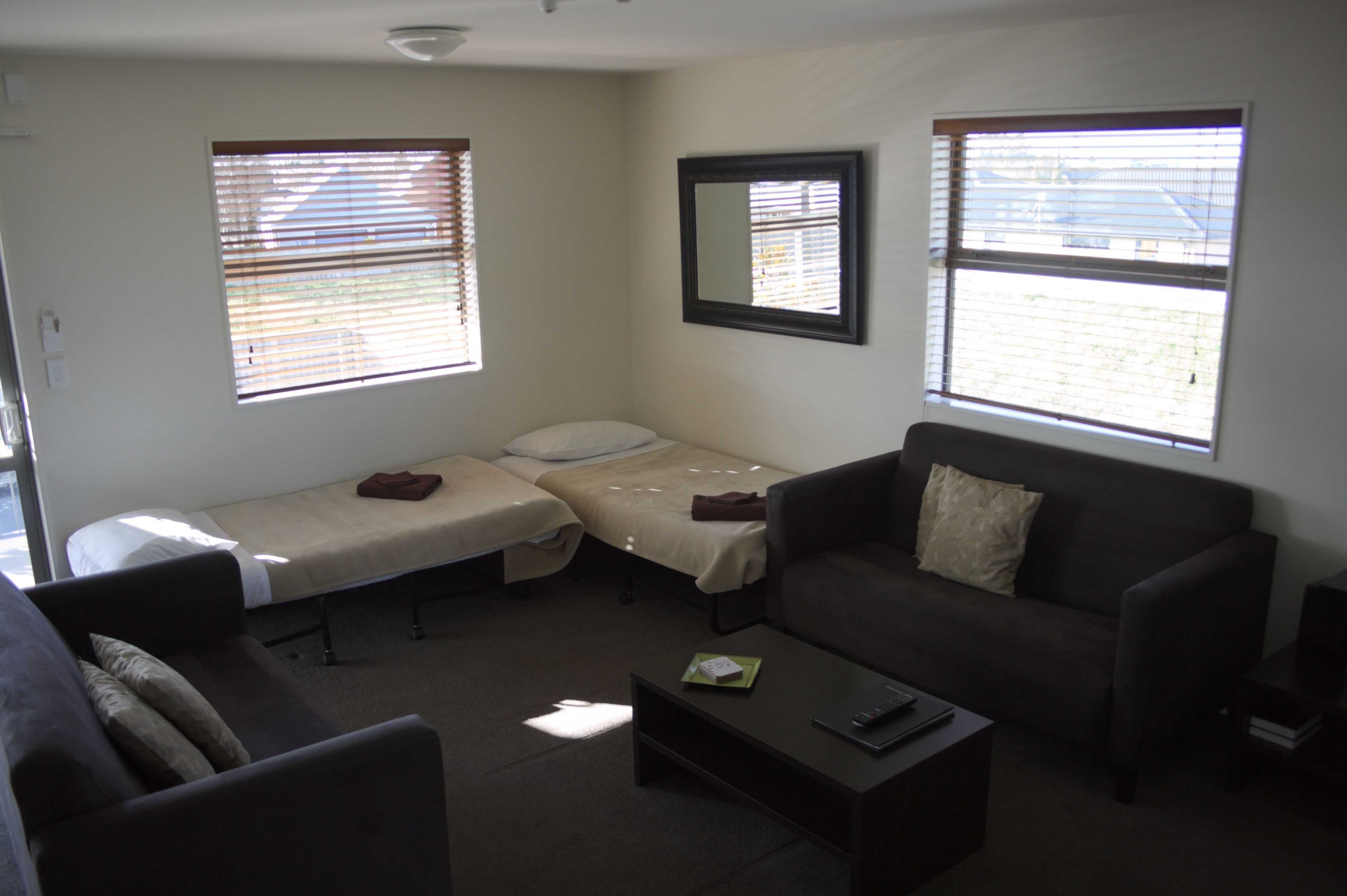 A small, furnished room with a sofa, a single bed, a coffee table, and two windows with blinds.