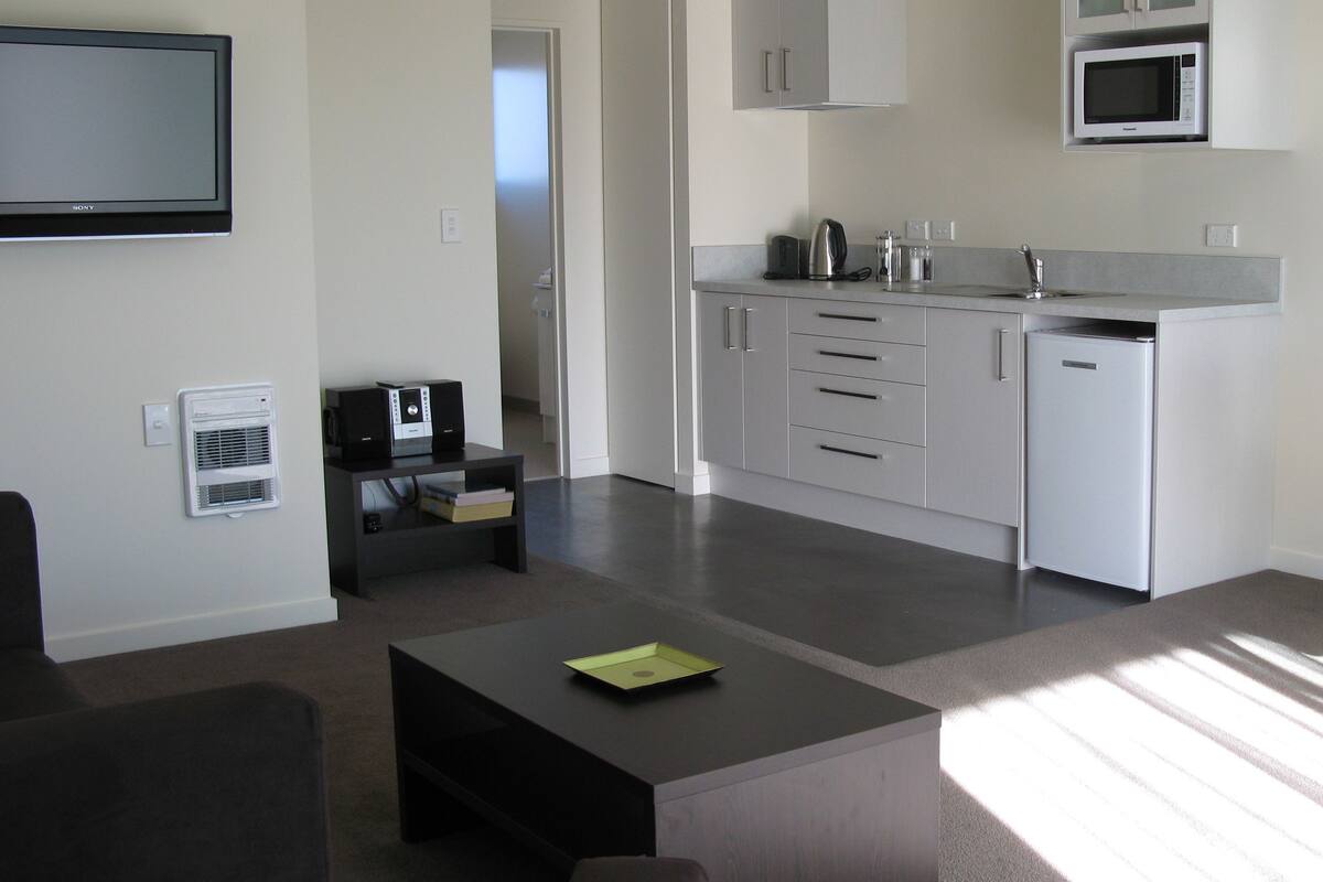 A modern kitchen with white cabinets, a built-in microwave, and a coffee maker. There's a small black table with a green folder on it, and a dark-colored sofa in the living area.