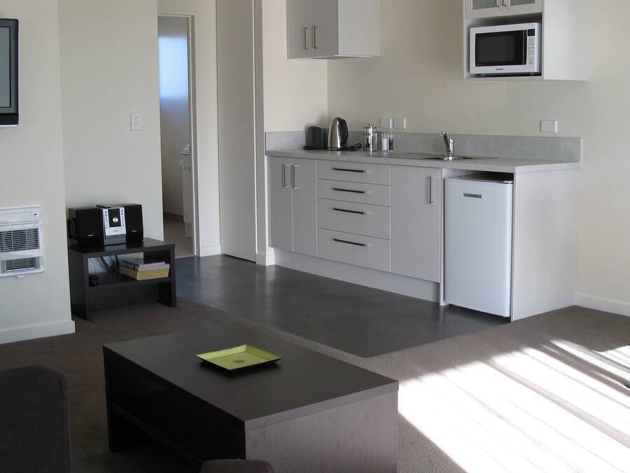 A modern kitchen with white cabinets, a built-in microwave, and a coffee maker. There's a small black table with a green folder on it, and a dark-colored sofa in the living area.