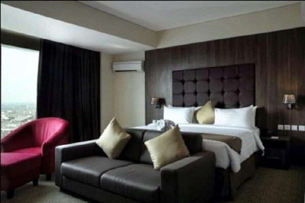 A hotel room with a large bed, a dark headboard, a red armchair, and a view of the city.