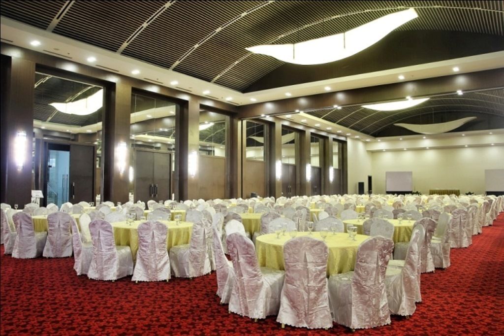 A large banquet hall with round tables covered in white tablecloths and chairs, a red carpet, and a modern ceiling design.