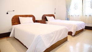 Une chambre d’hôtel avec deux lits simples, des têtes de lit en bois, du linge de lit blanc, une table de chevet avec une lampe et une fenêtre avec des rideaux légers.