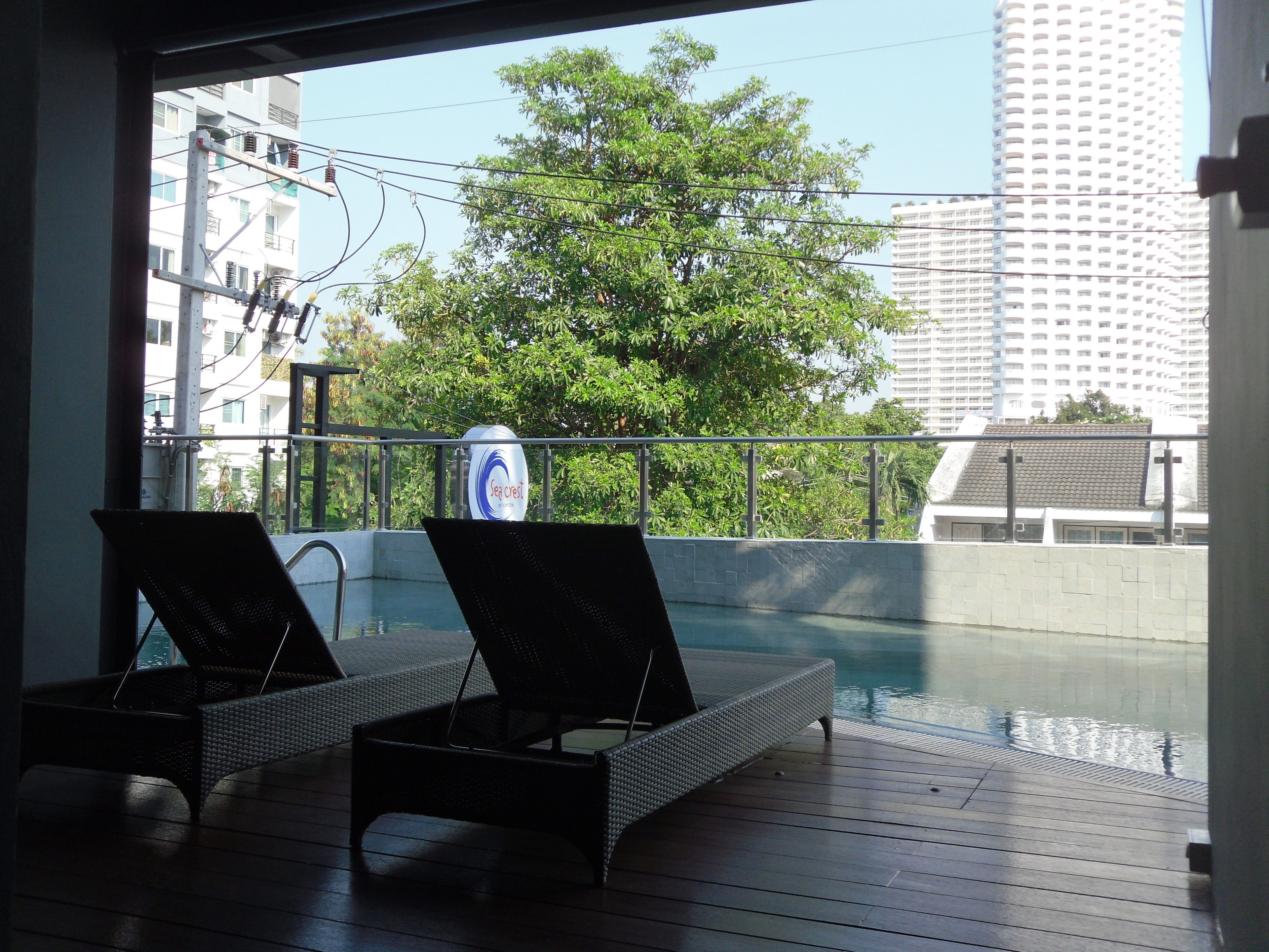 A balcony with two lounge chairs, a pool, and a view of a tree and buildings.