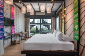 Quarto de hotel moderno com cama grande, televisão, escrivaninha com cadeira e vista da cidade através de grandes janelas.