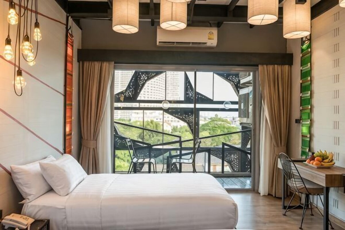 A hotel room with a bed, a desk, a chair, and a view of greenery through large windows.