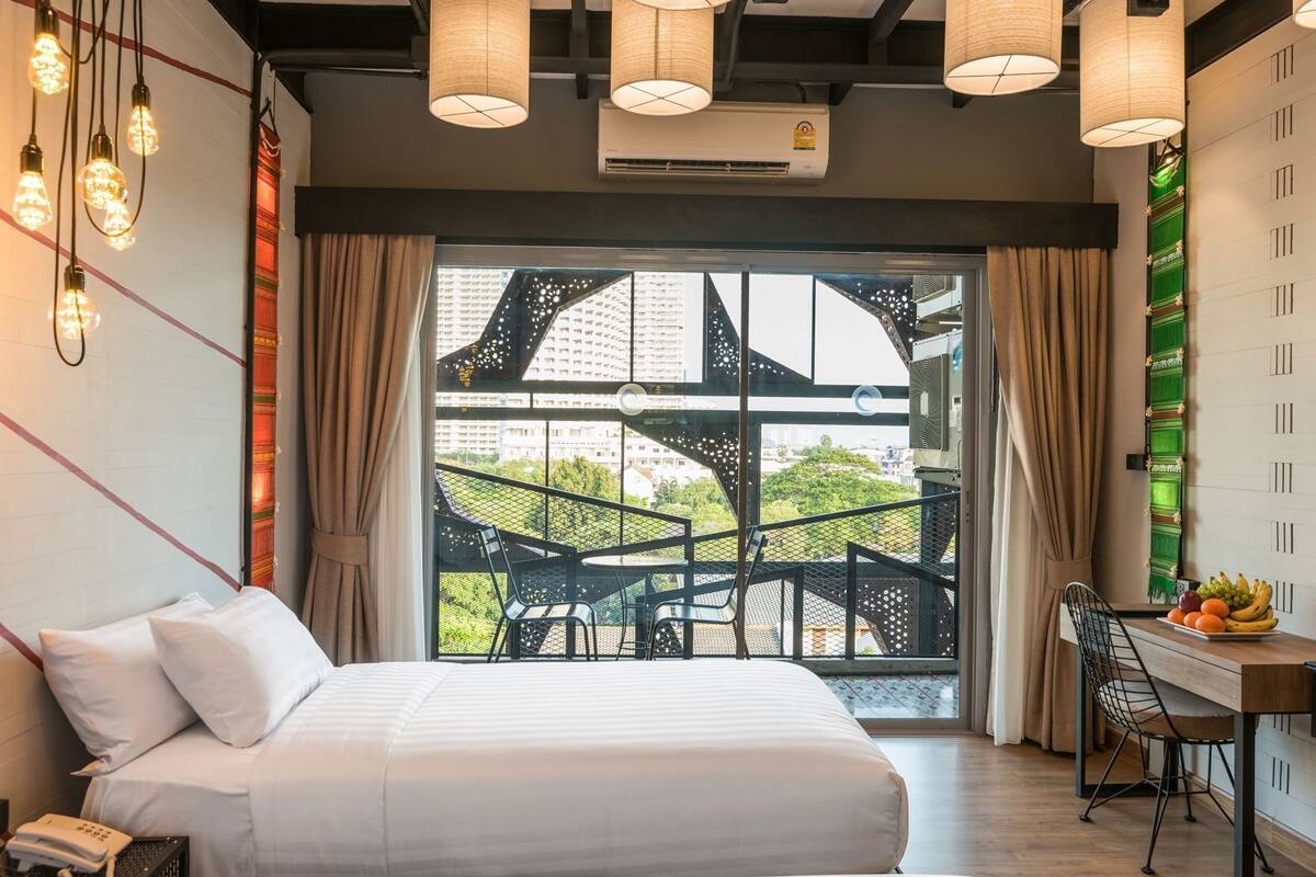 A hotel room with a bed, a desk with fruit, a view of buildings, and hanging lights.