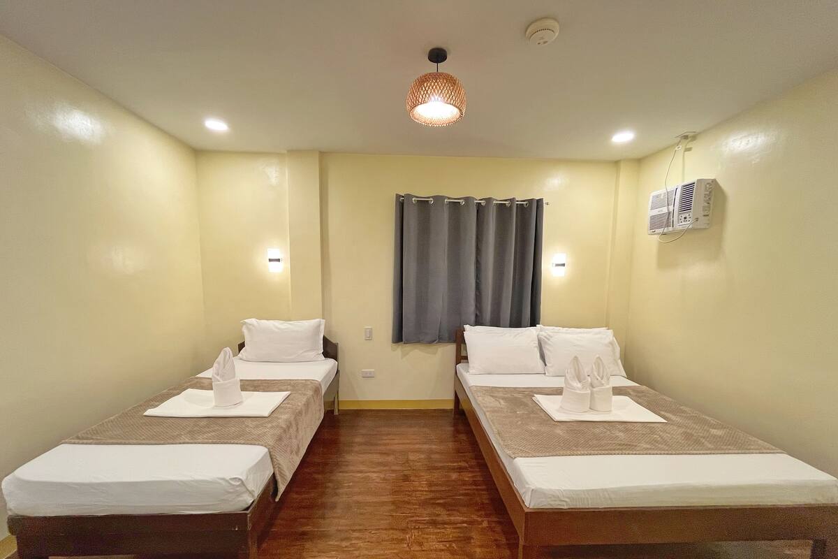 Habitación de hotel con dos camas individuales, suelo de madera, una lámpara en el techo y una ventana con cortinas.