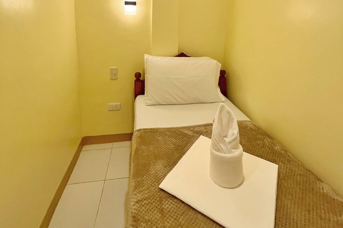 Una habitación de hotel pequeña y estrecha con una cama individual, una mesita de noche y una lámpara fijada a la pared.