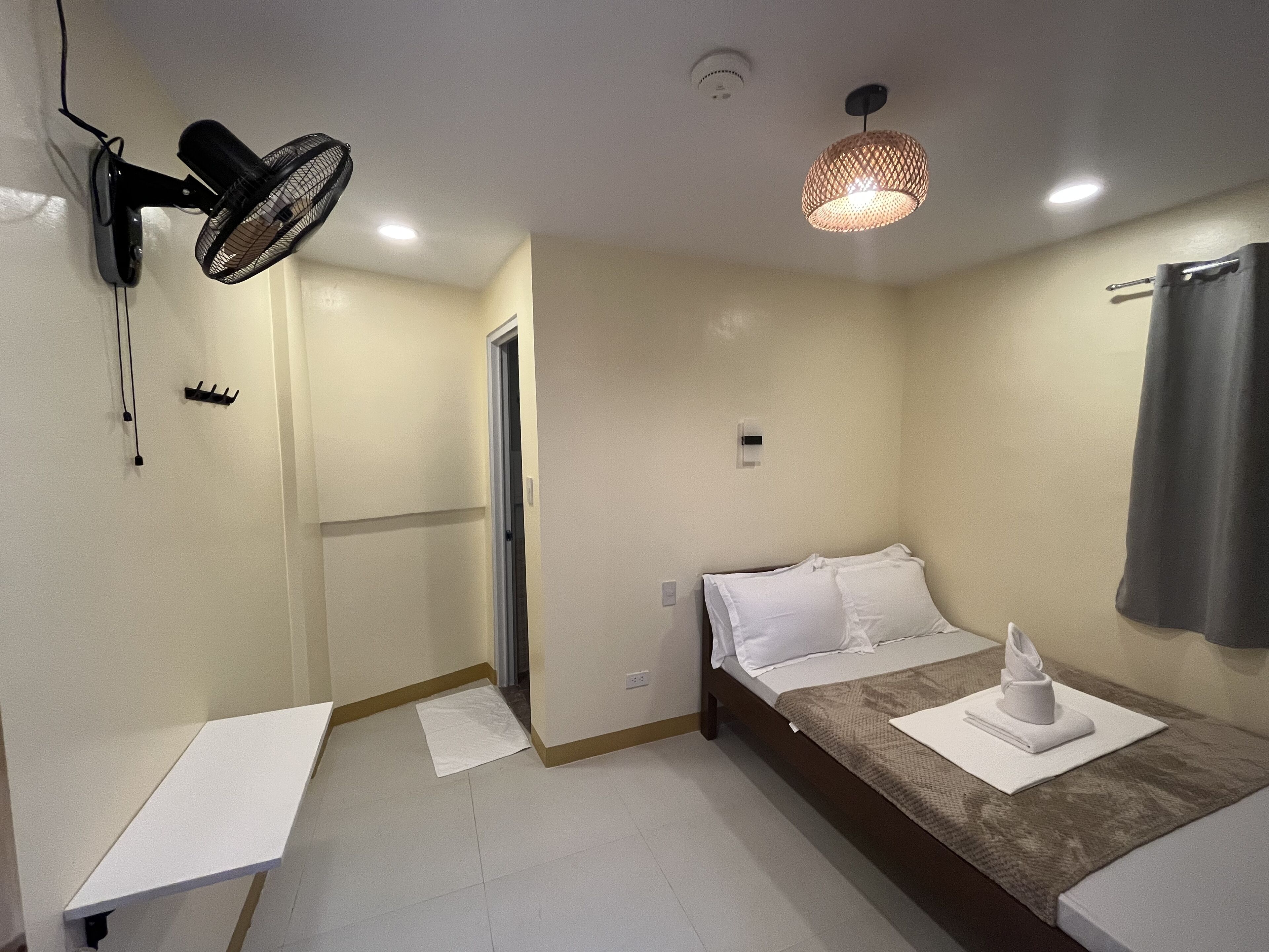 Quarto pequeno e bem organizado, com uma cama de solteiro, mesa de cabeceira, ventoinha na parede, luminária suspensa e uma porta que dá para outro cômodo.