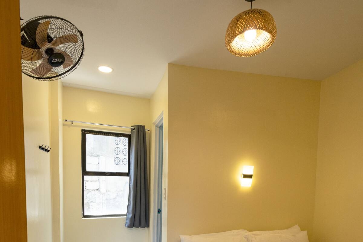Habitación de hotel con cama, escritorio, silla, ventana con cortinas, ventilador de pared, luz de techo e iluminación en la pared.
