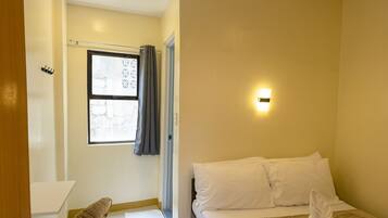 A hotel room with a bed, a desk, a chair, a window with curtains, a wall-mounted fan, a ceiling light, and a wall-mounted light fixture.