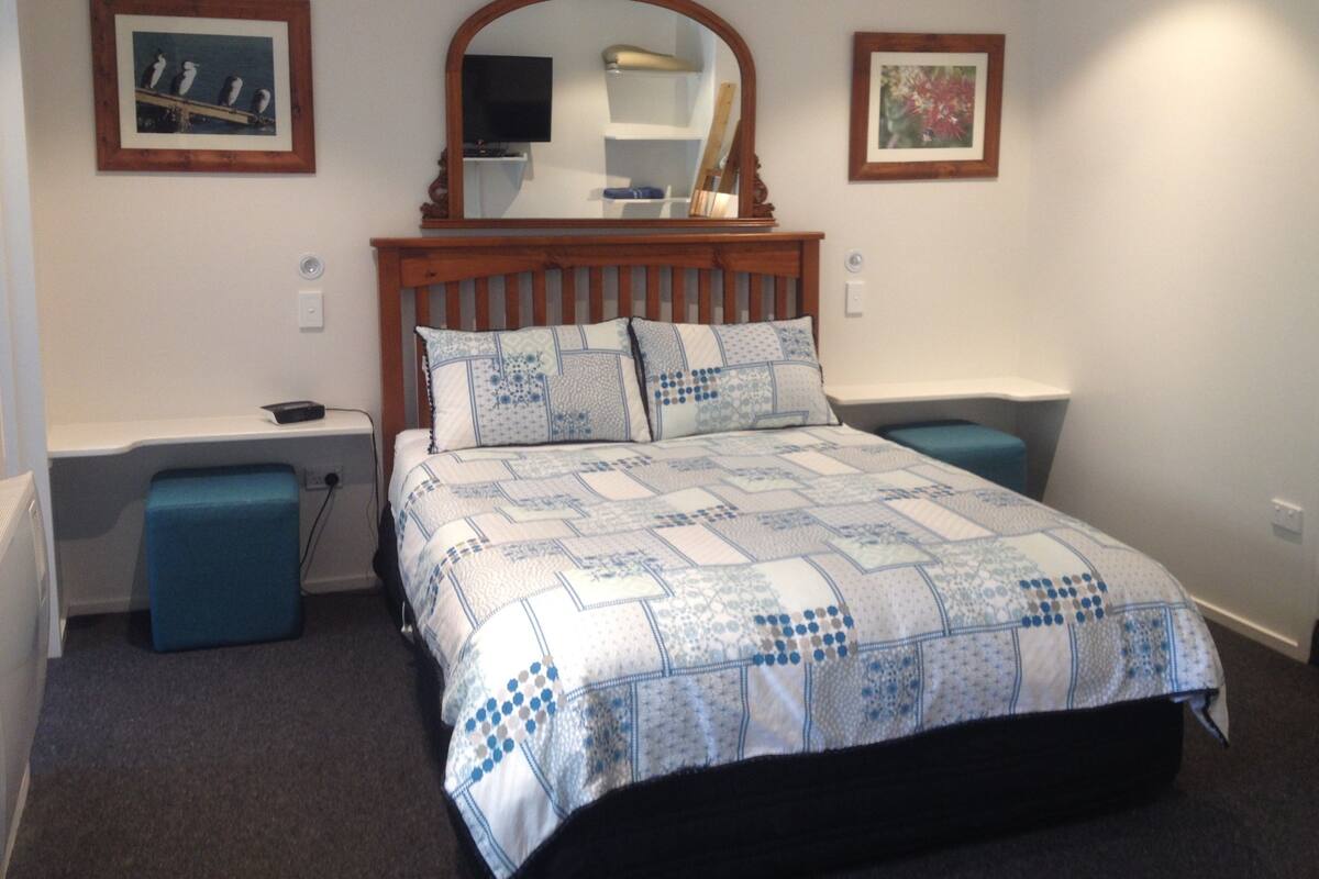 A bedroom with a wooden headboard, a bed with a quilt, two bedside tables, a nightstand, a mirror, and framed pictures on the walls.