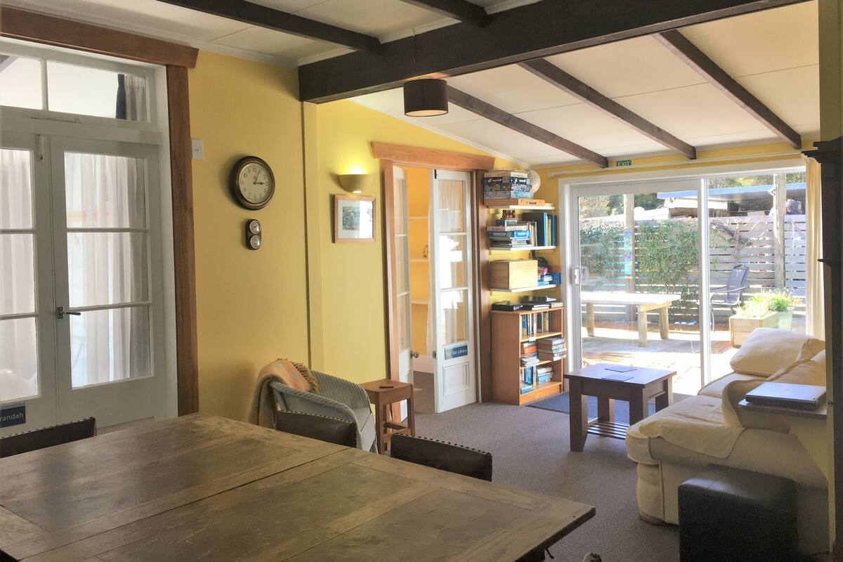 A cozy room with a wooden table, a sofa, and a bookshelf. There is a door leading to a balcony with a view of the outside.