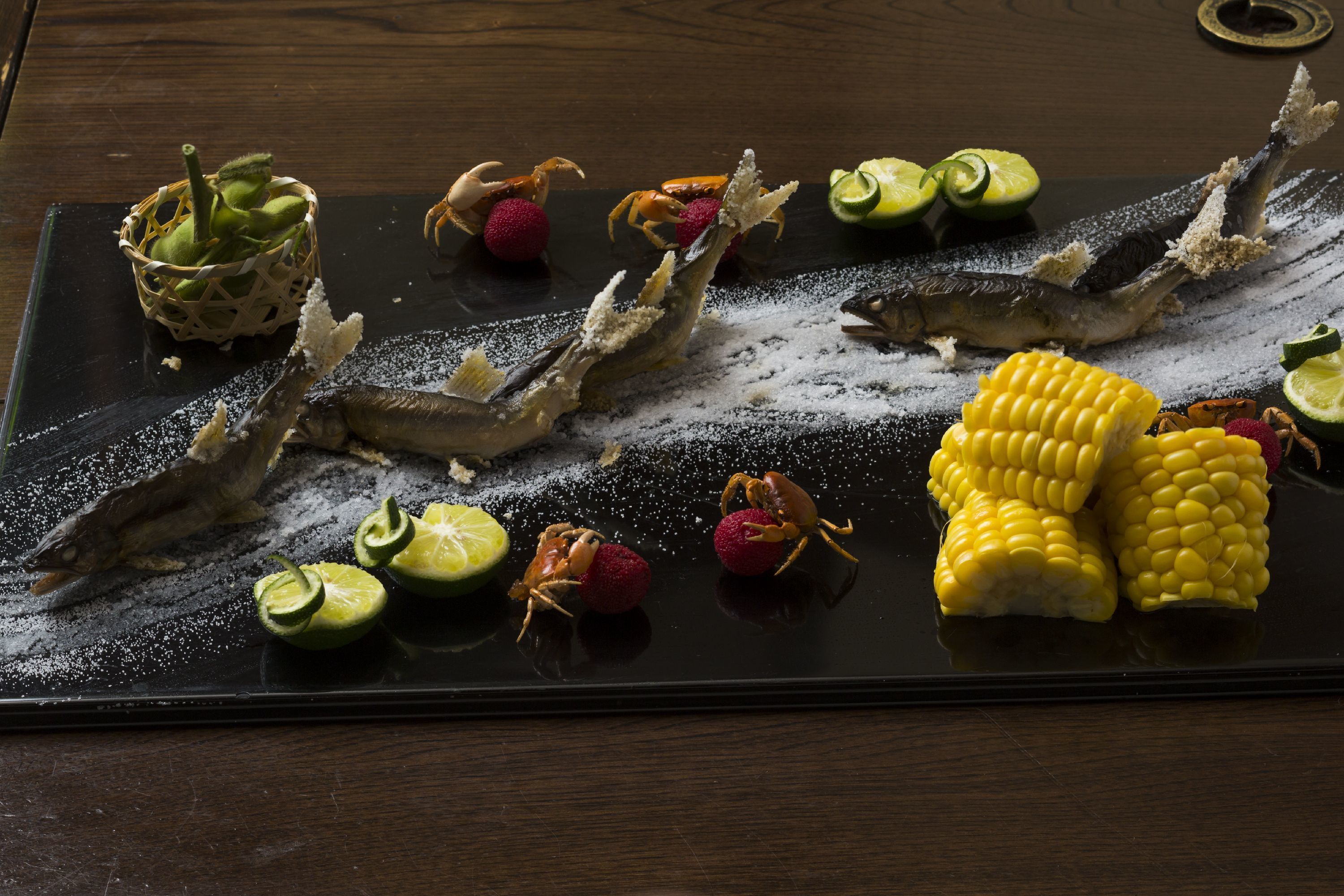 A black slate serving tray with various seafood and vegetables, including fish, corn, and lime slices.