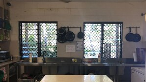 A kitchen with a large table, two windows with metal grilles, and various kitchen utensils and appliances. - Frogshollow Backpackers (Darwin)