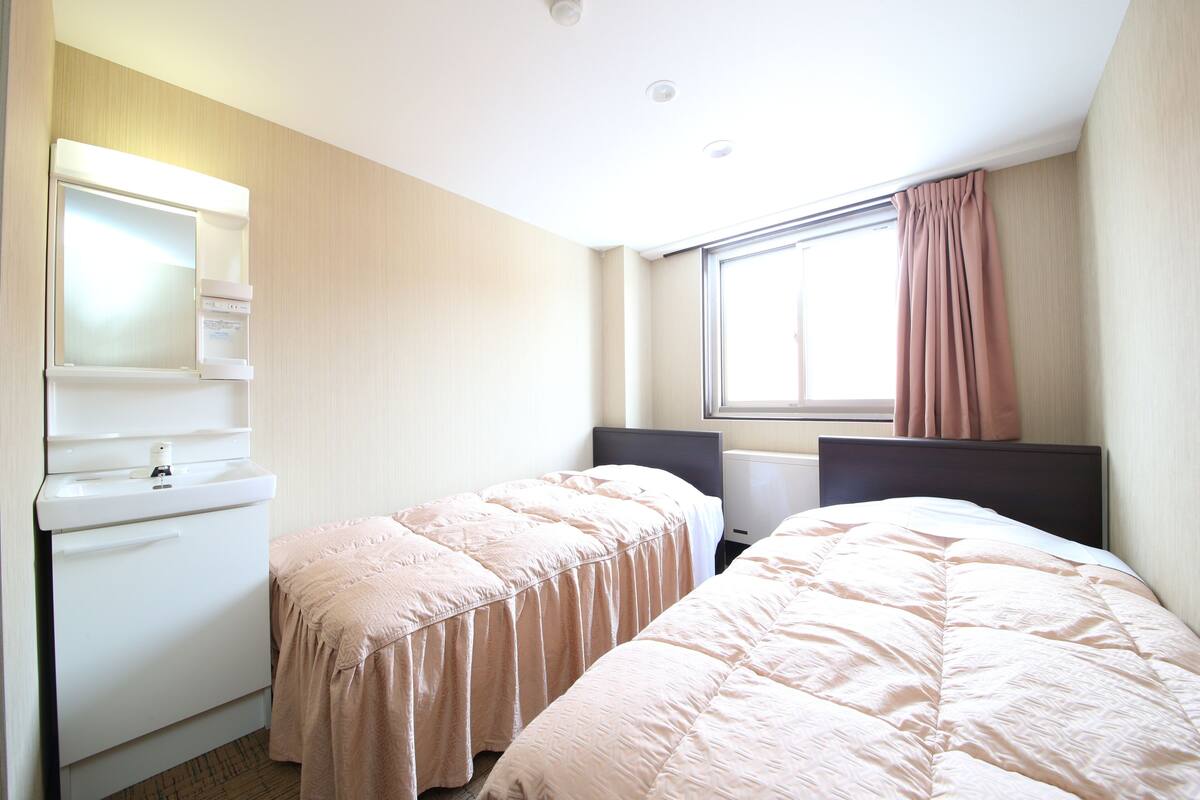 A hotel room with two beds, a small kitchenette, a window with curtains, and a ceiling light.