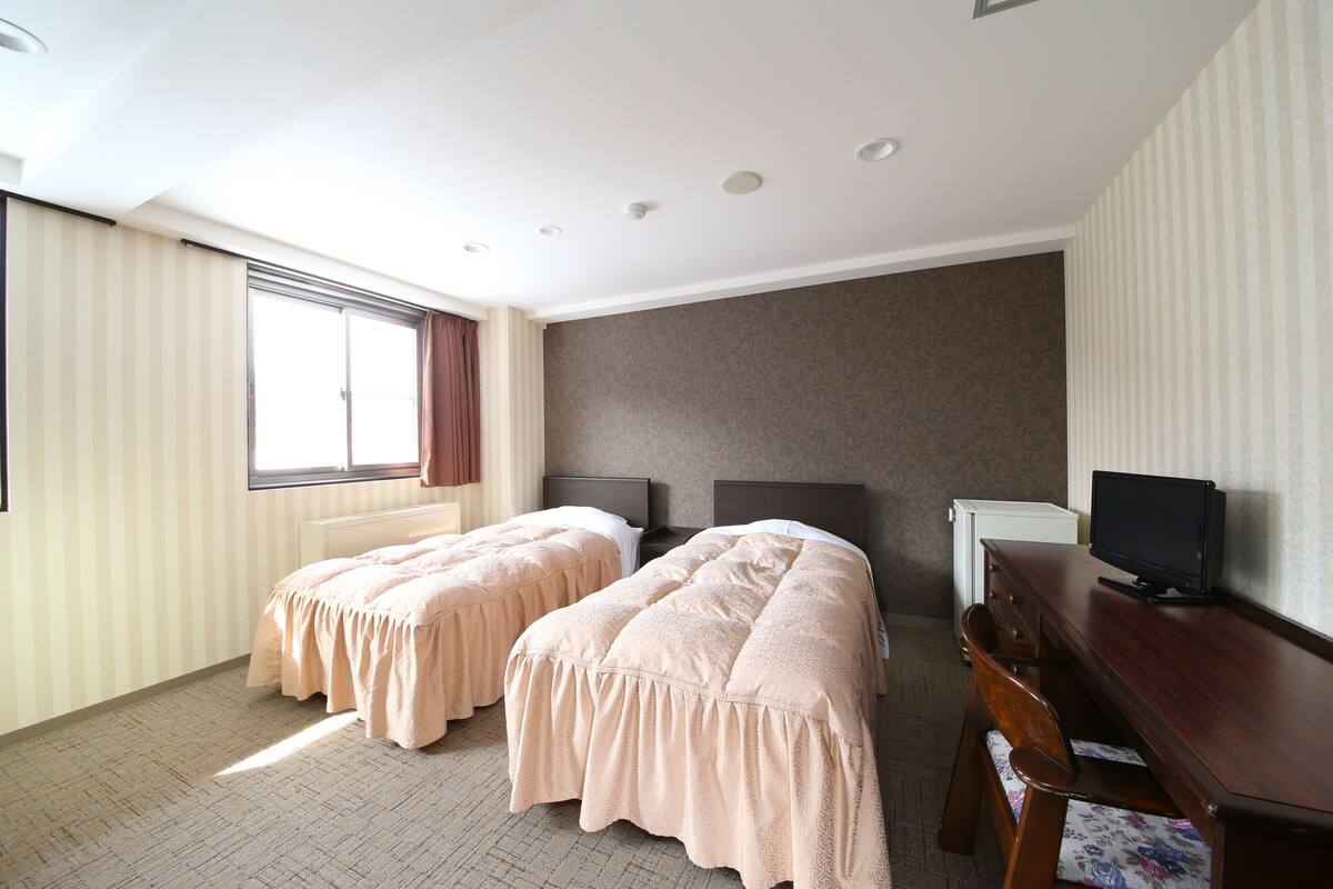 A hotel room with two beds, a desk, a television, and a window with curtains.