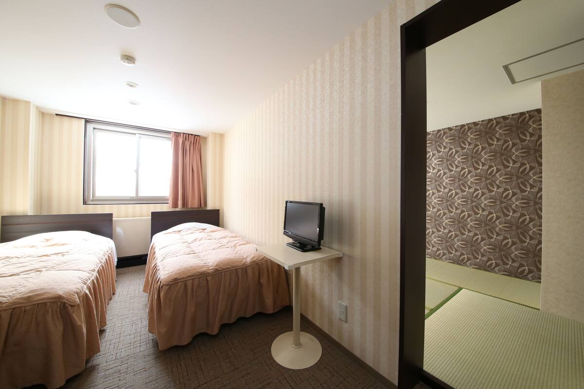 A hotel room with two beds, a small desk with a television, a window with curtains, and patterned wallpaper.