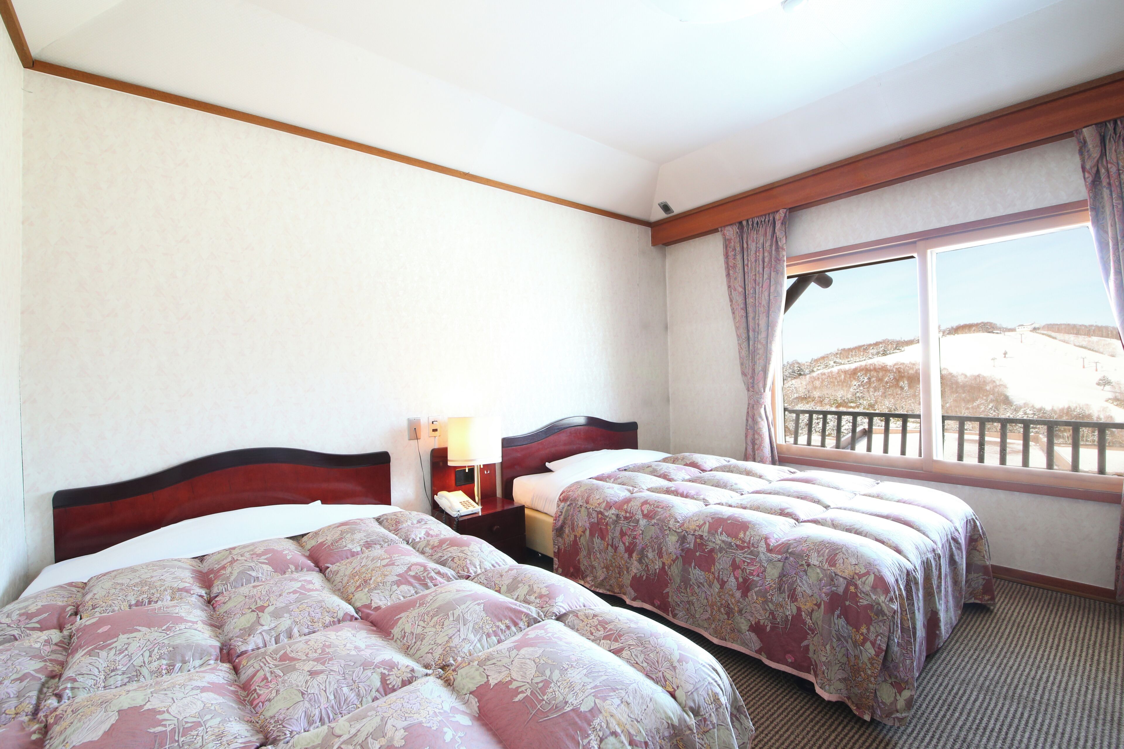 Ein Hotelzimmer mit zwei Betten, einem Nachttisch, einer Lampe und einem Fenster mit Blick auf eine hügelige Landschaft.