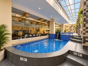 Una piscina interior con techo de vidrio, rodeada por un restaurante con mesas visibles y una cascada.