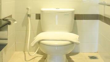 Inodoro blanco con botón de descarga y un accesorio para bidé en un baño con azulejos blancos y grises.