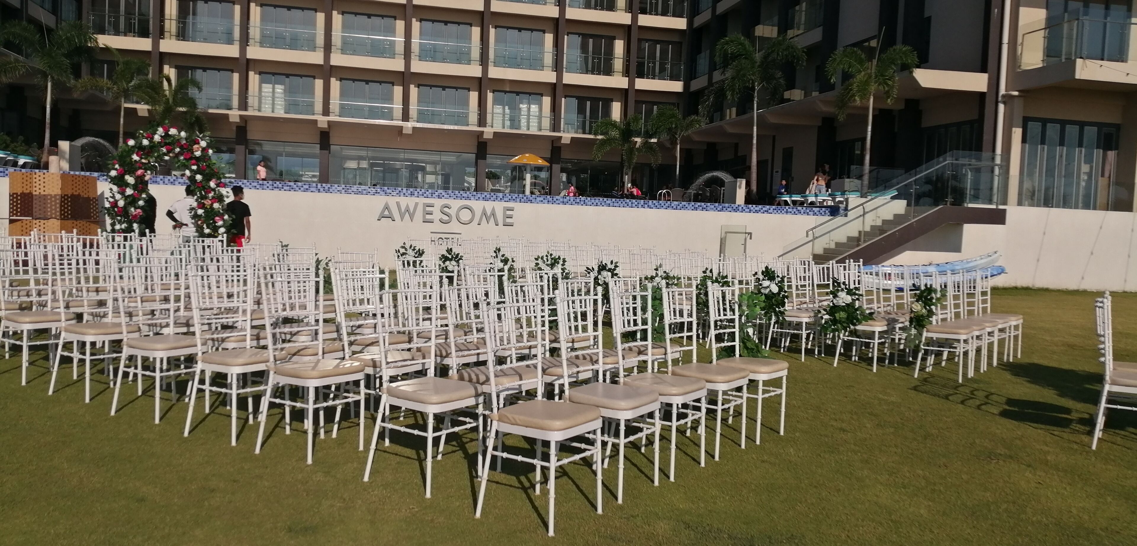 Un hotel llamado "AWESOME" con una terraza acondicionada para eventos, que presenta filas de sillas blancas con asientos y respaldos acolchados, dispuestas sobre un césped.