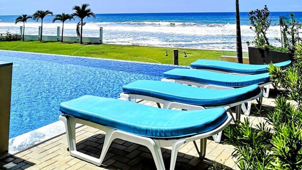 Un área junto a la piscina con sillones de descanso, una palmera y vista al océano.