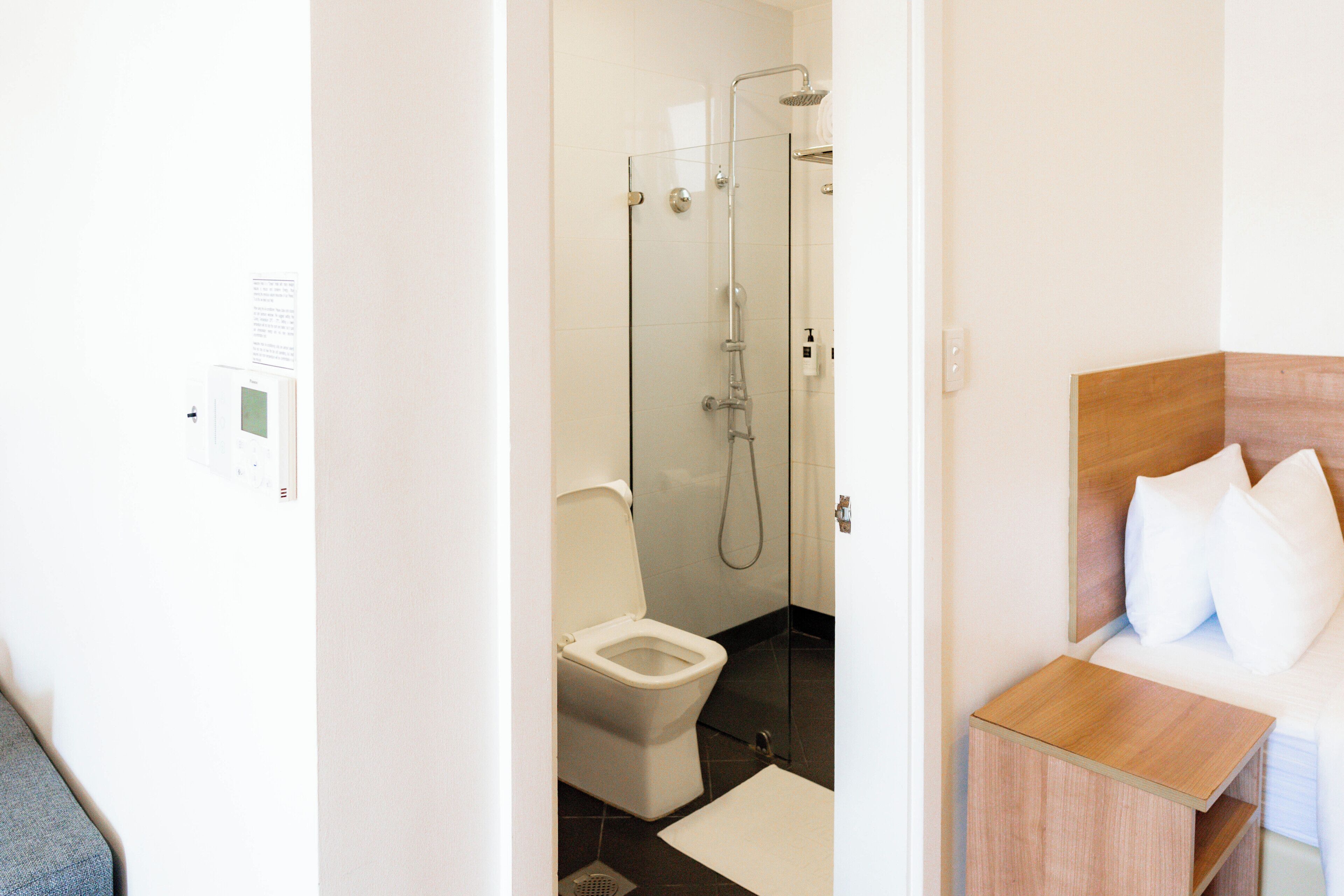A hotel bathroom with a toilet, shower, and a bed with pillows.