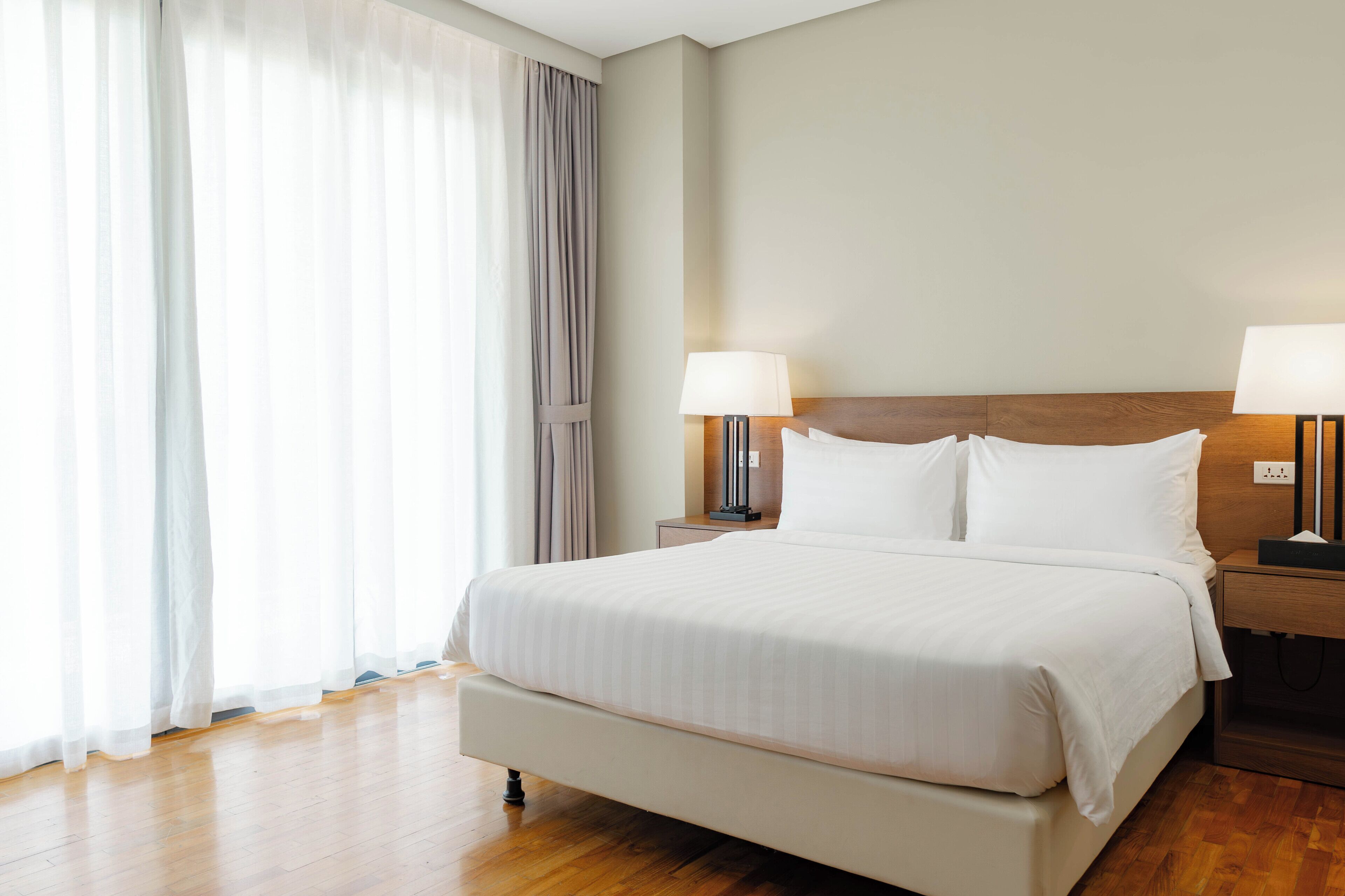 Une chambre d’hôtel avec un grand lit, deux tables de chevet avec des lampes, une tête de lit en bois et une fenêtre avec des rideaux légers.