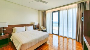 Habitación de hotel con una cama grande, ventilador de techo, mesita de noche con lámpara, una silla y vista a la playa desde las ventanas.