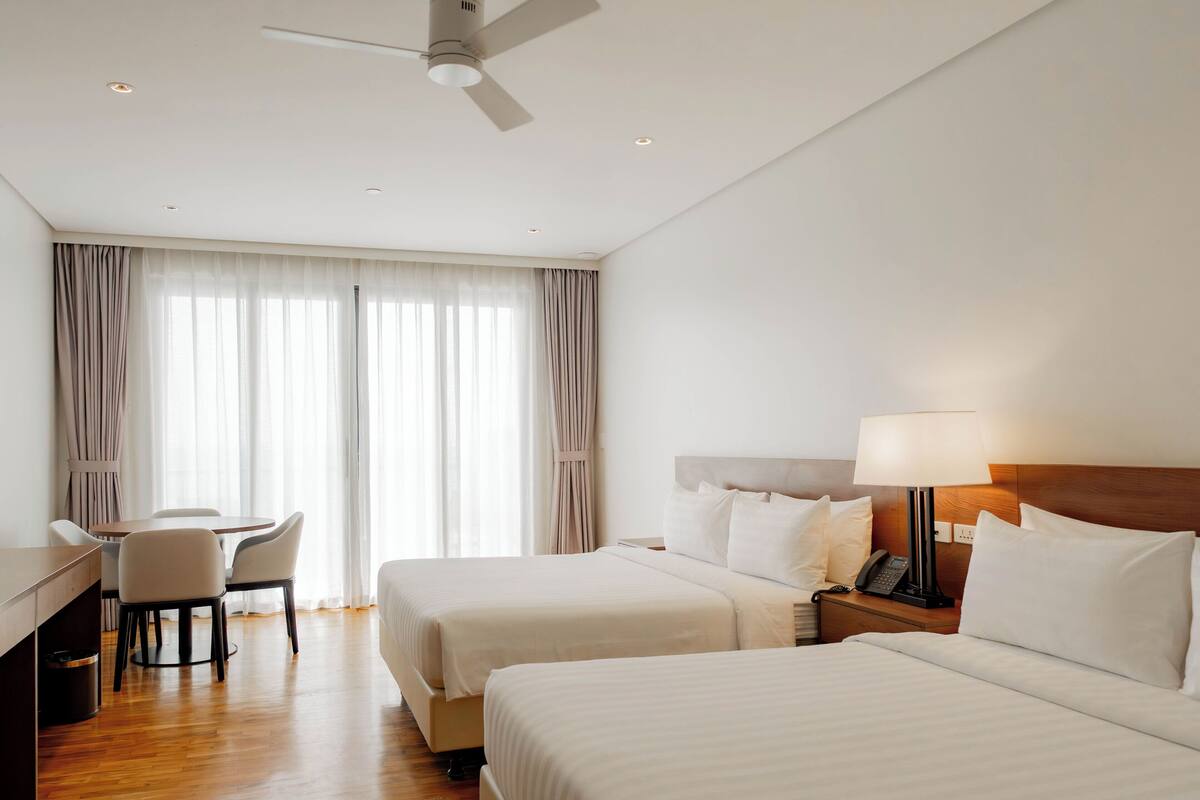 Une chambre d’hôtel avec deux lits, un bureau, une chaise, une lampe et un ventilateur de plafond.