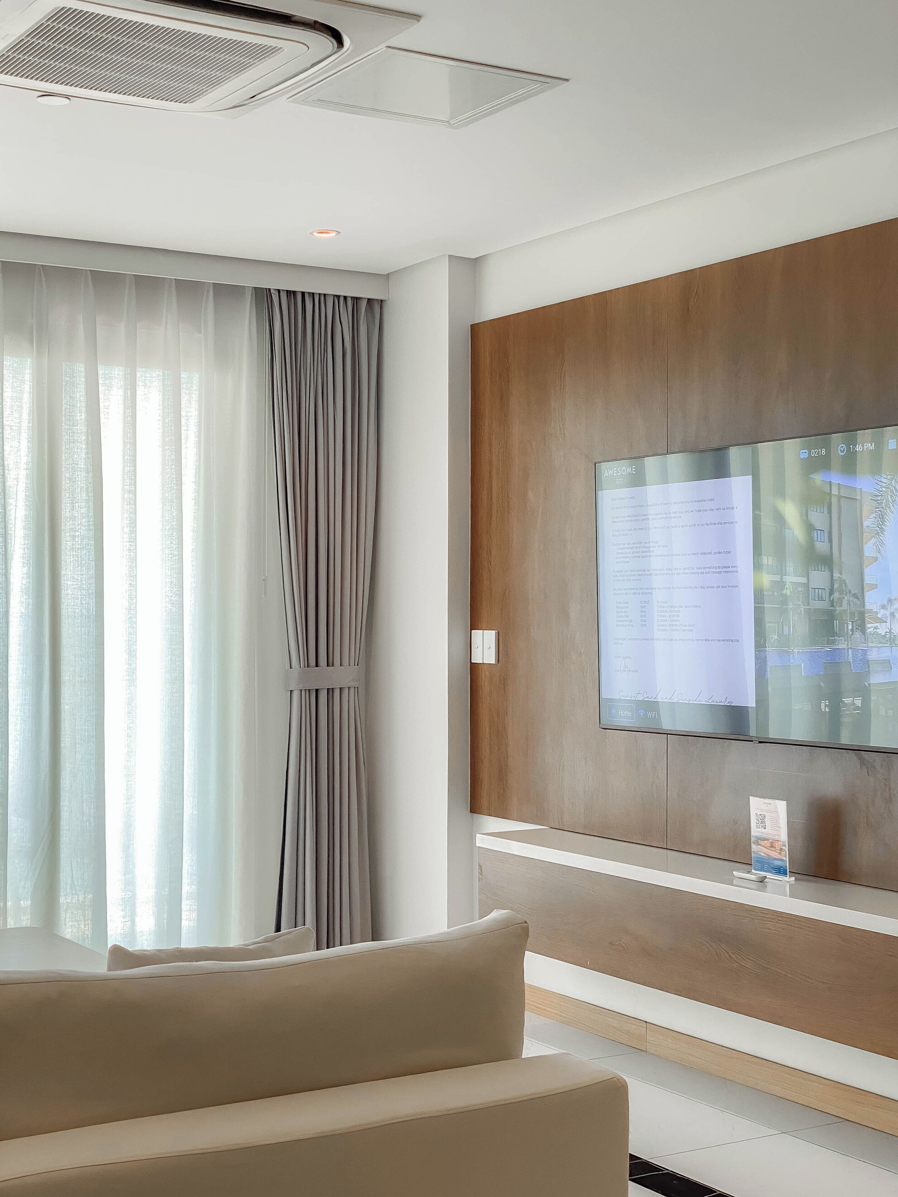 A modern hotel room with a large flat-screen TV mounted on a wooden panel, a beige sofa, and sheer curtains on the window.