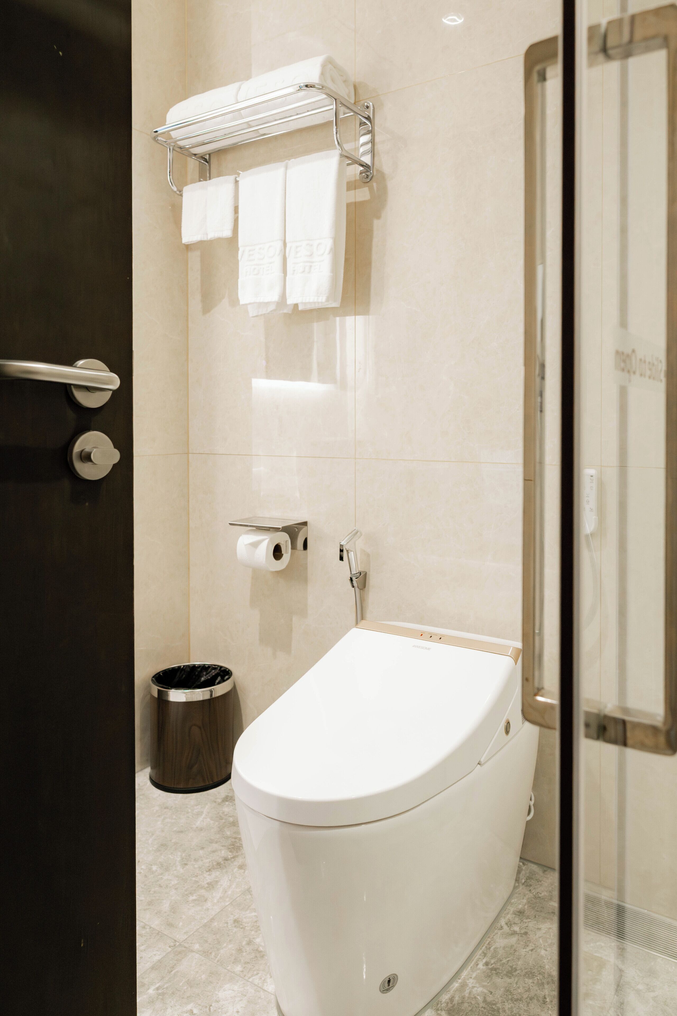 A bathroom with a toilet, a towel rack with towels, a trash bin, and a shower area.