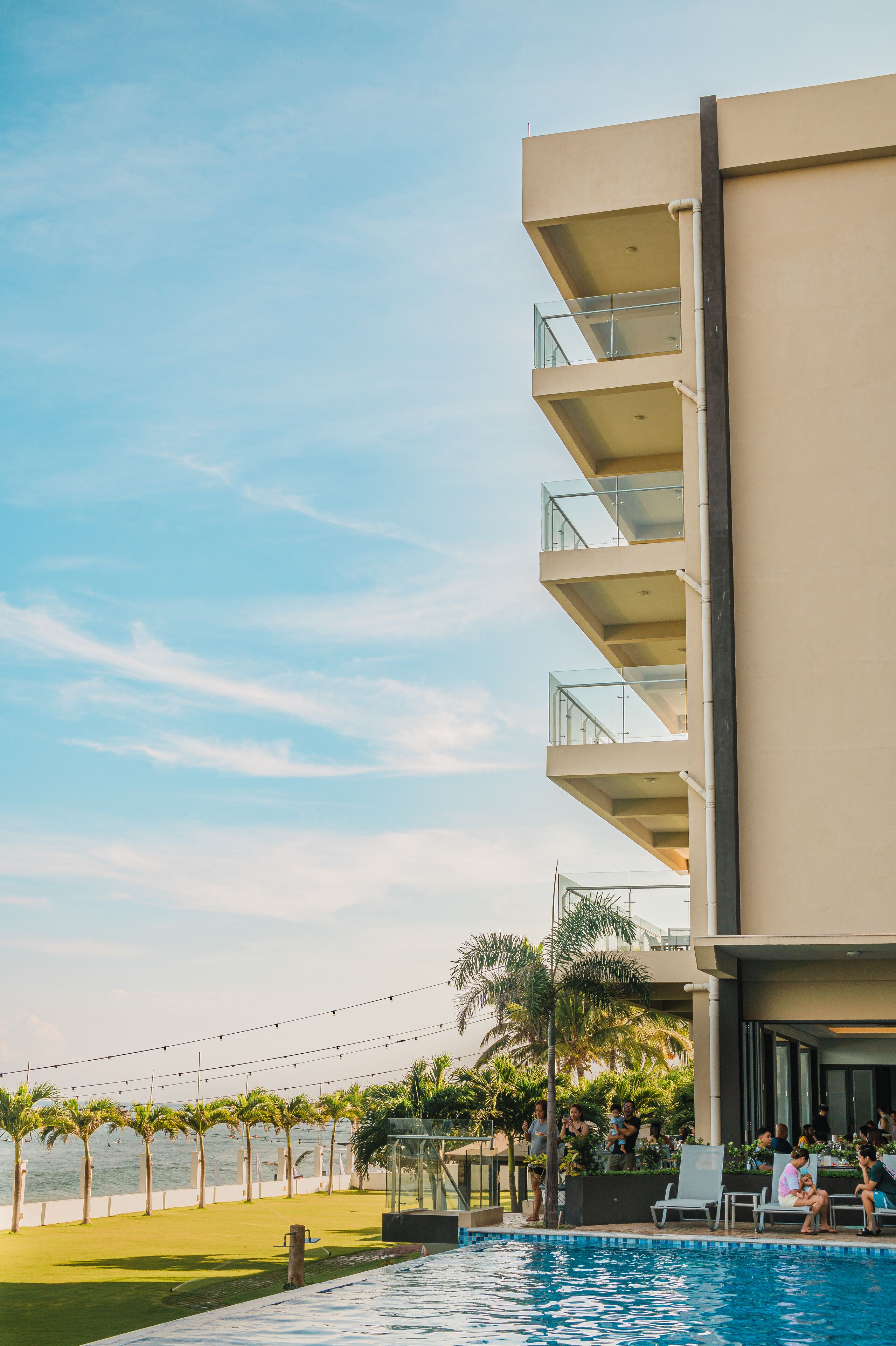 A poolside area with a clear view of the ocean, a building with multiple balconies, and a grassy area with palm trees.