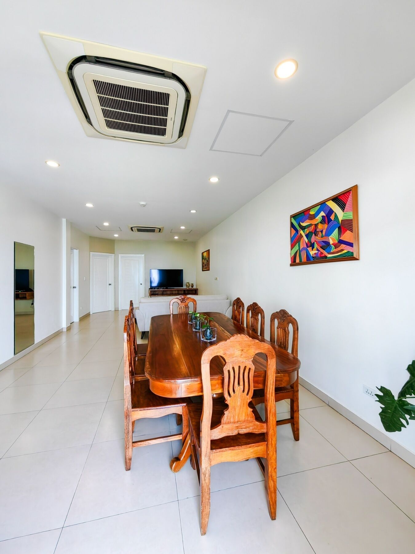 A spacious dining area with a large wooden table and chairs, a ceiling-mounted air conditioning unit, recessed lighting, and a colorful abstract painting on the wall.