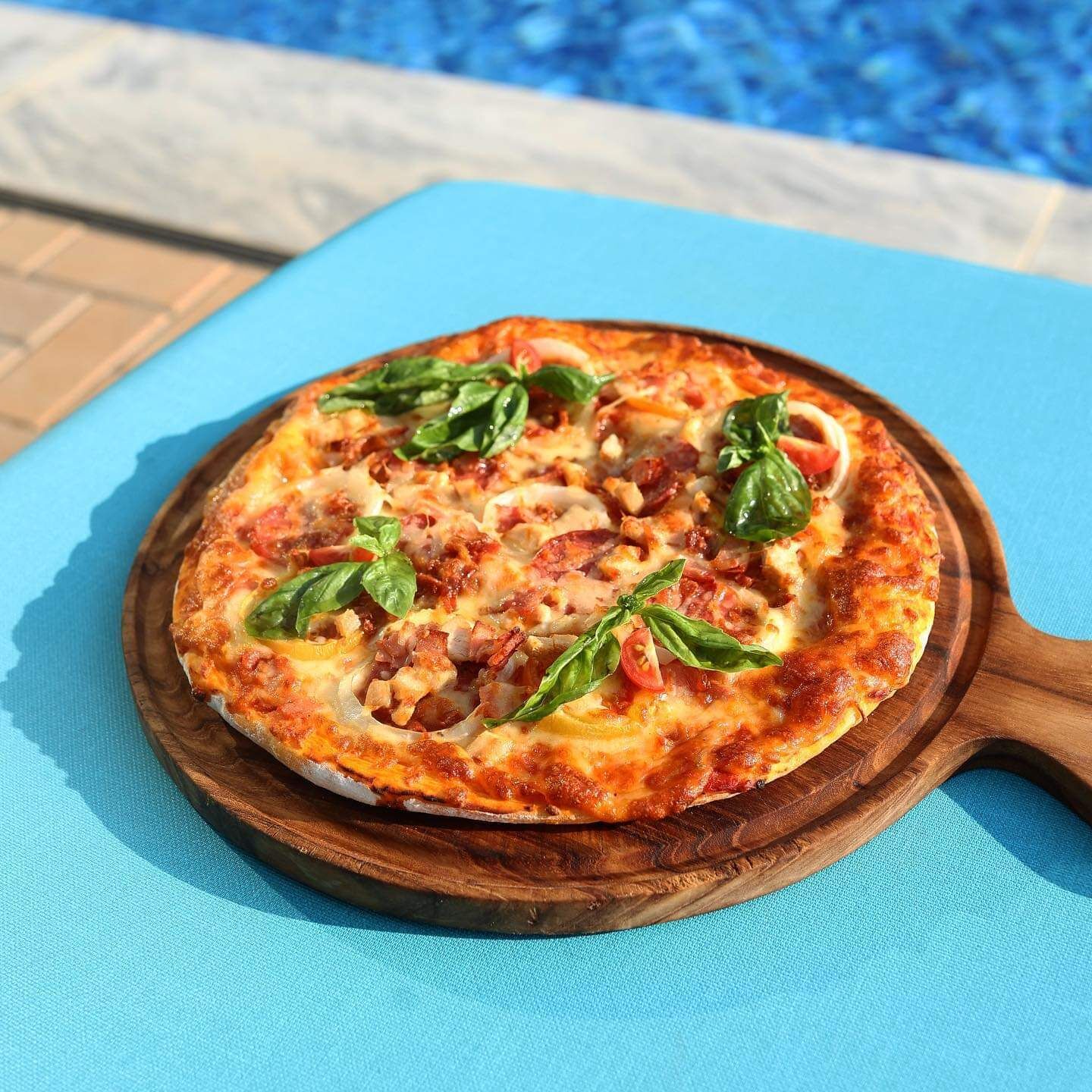 Une pizza sur une planche en bois, garnie de feuilles de basilic, posée sur un tissu bleu près d’une piscine.