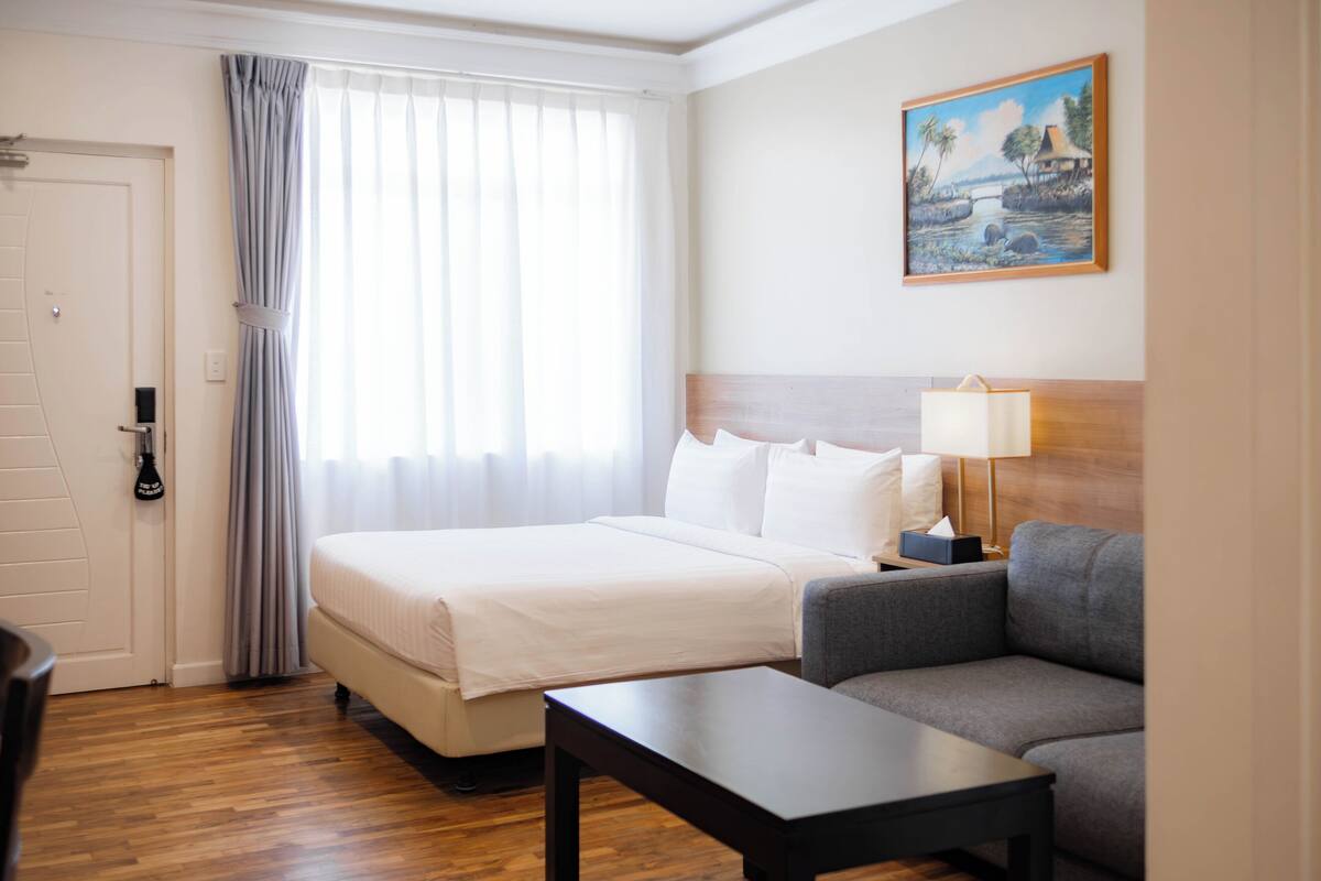 Une chambre d’hôtel avec un lit, un canapé, une table, un tableau au mur et une fenêtre avec des rideaux.