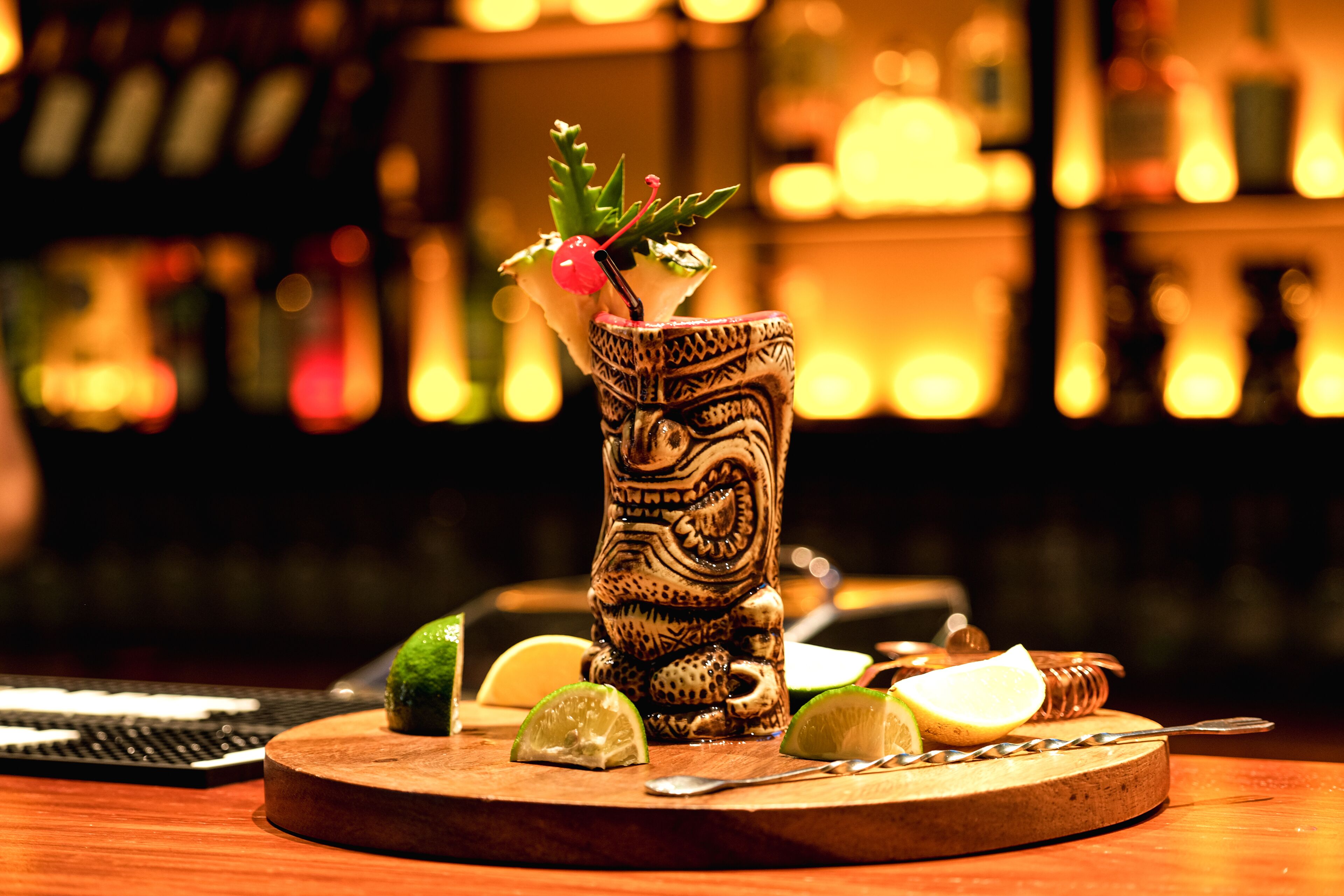 A cocktail in a carved wooden glass on a wooden board with lime wedges and a spoon.