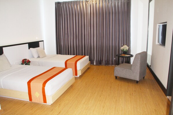 A hotel room with two beds, a small table with a vase of flowers, a grey armchair, and a TV mounted on the wall.