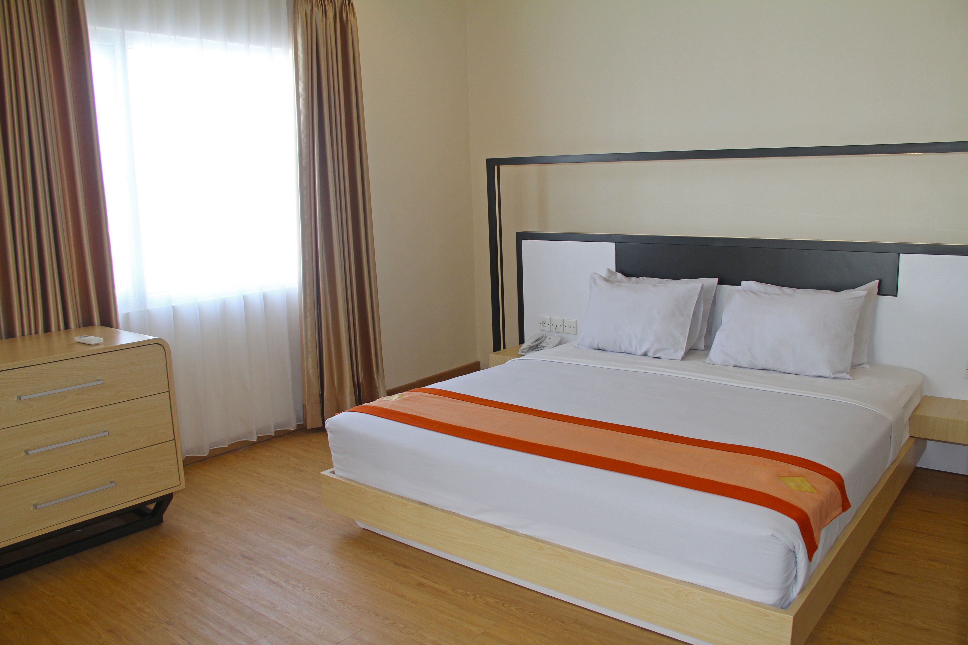 A hotel room with a bed, wooden dresser, and a window with curtains.