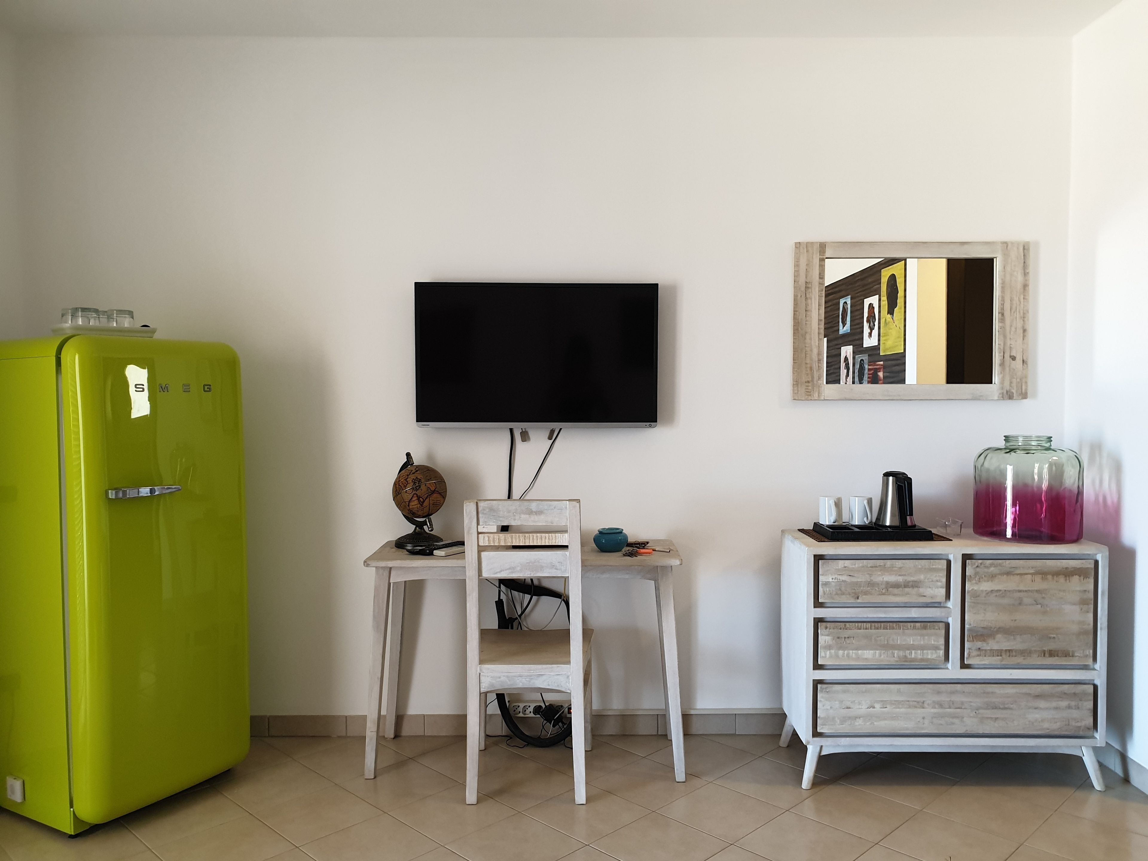 Una habitación con un refrigerador verde, un escritorio blanco con un globo terráqueo, un televisor de pantalla plana, un armario de madera y un espejo en la pared.