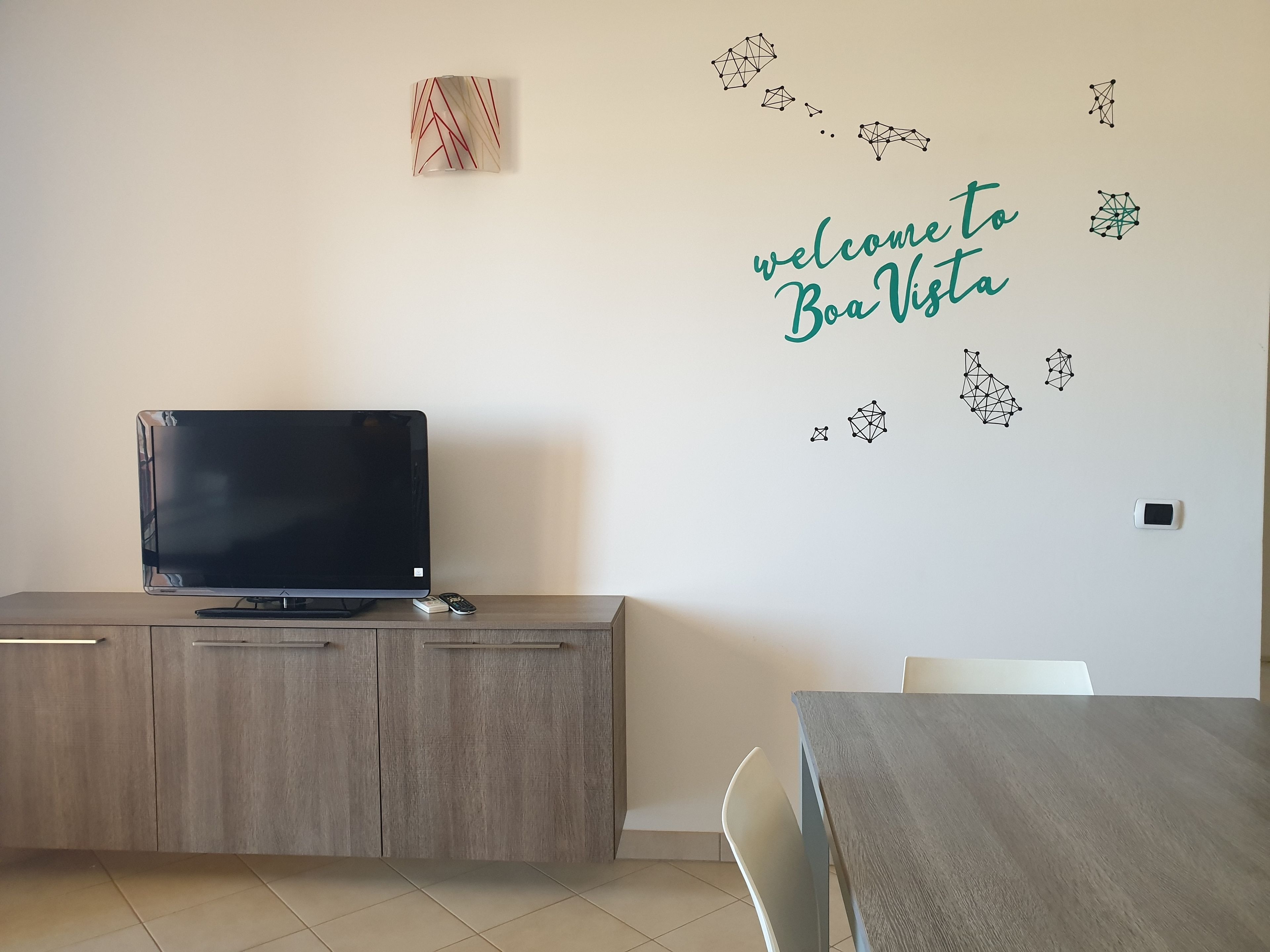 A modern living room with a flat-screen TV on a wooden cabinet, a dining table with chairs, and a wall with a 'welcome to Boa Vista' sign and geometric wall art.