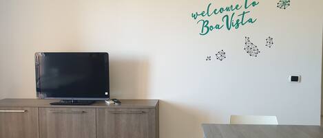A modern living room with a flat-screen TV on a wooden cabinet, a dining table with chairs, and a wall with a 'welcome to Boa Vista' sign and geometric wall art.