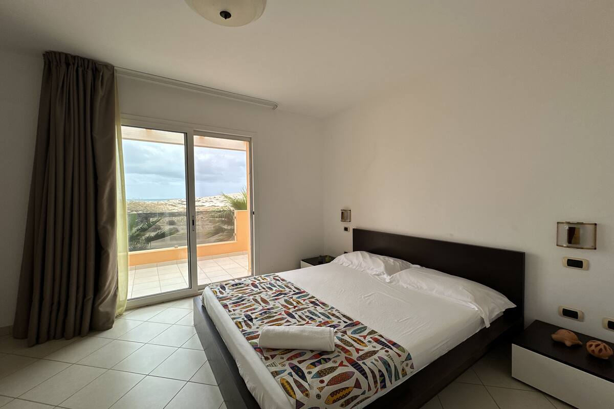 Ein Schlafzimmer mit Bett, Nachttisch, einem Fenster mit Vorhängen und Blick ins Freie.