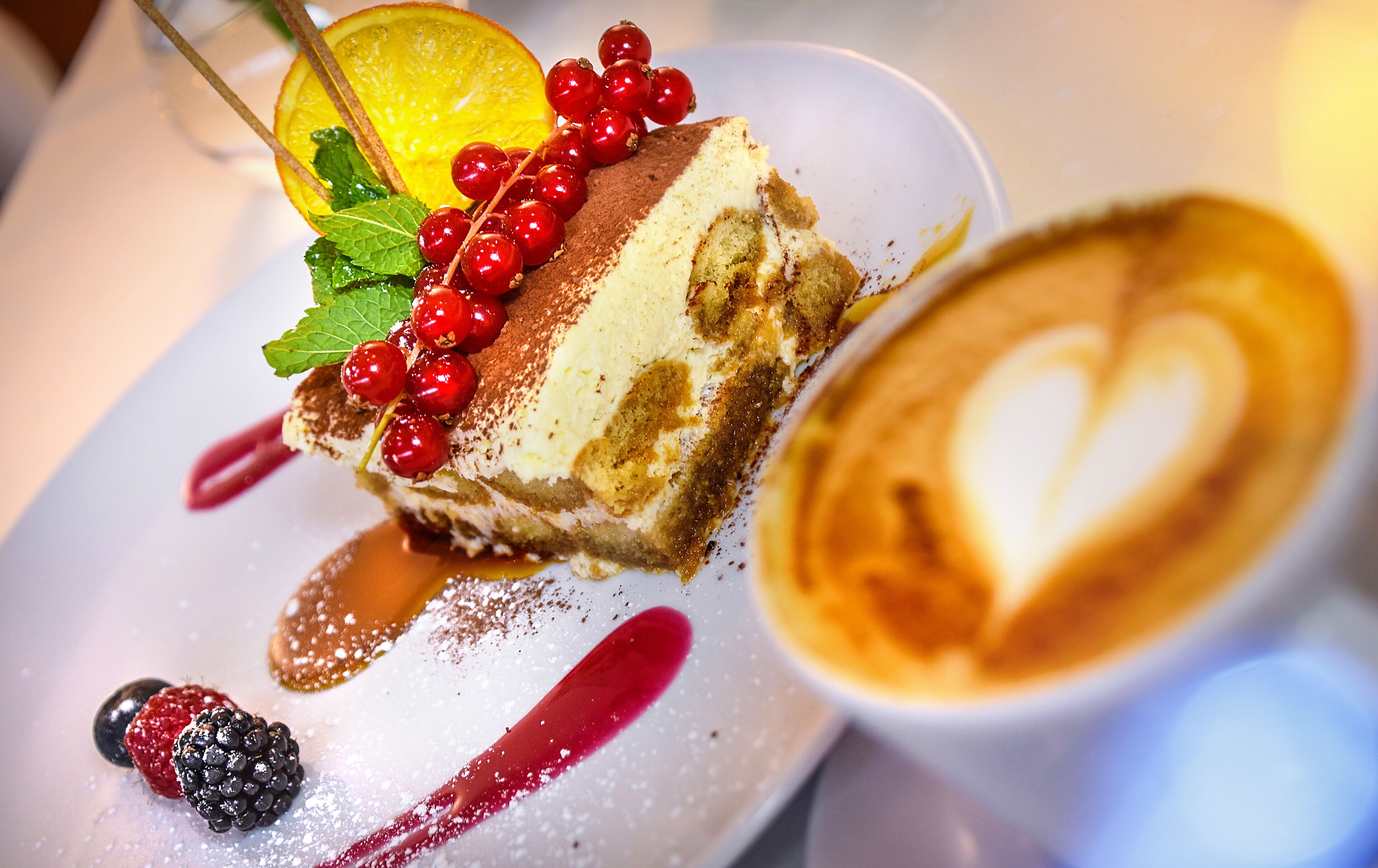 A dessert plate with a slice of cake, berries, and a heart-shaped coffee drink.