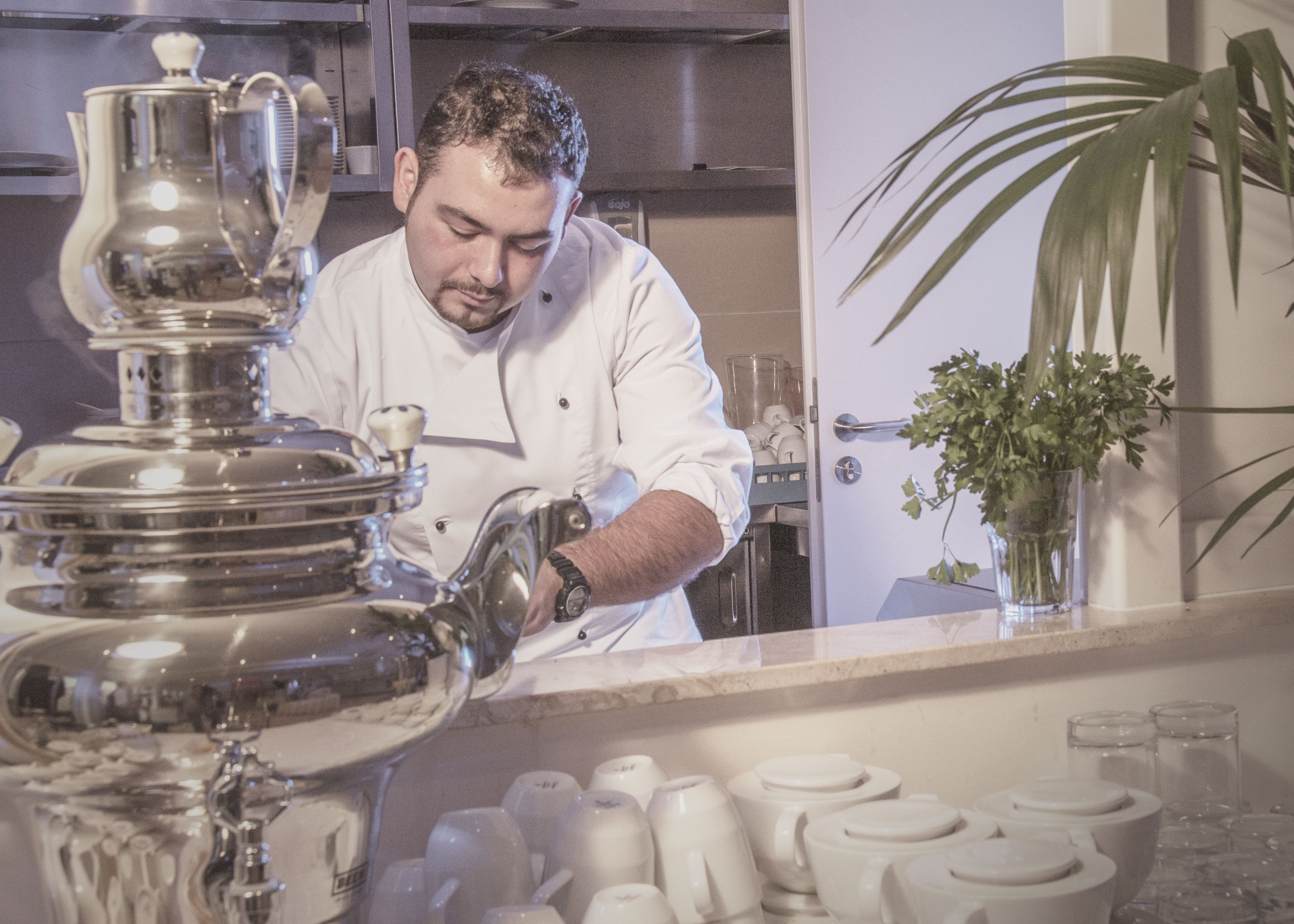 A chef in a white uniform preparing food in a kitchen with stainless steel appliances and a potted plant.