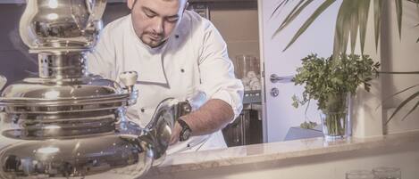 A chef in a white uniform preparing food in a kitchen with stainless steel appliances and a potted plant.
