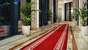Un couloir d’hôtel avec un tapis rouge, des plantes en pot et un sol en marbre.